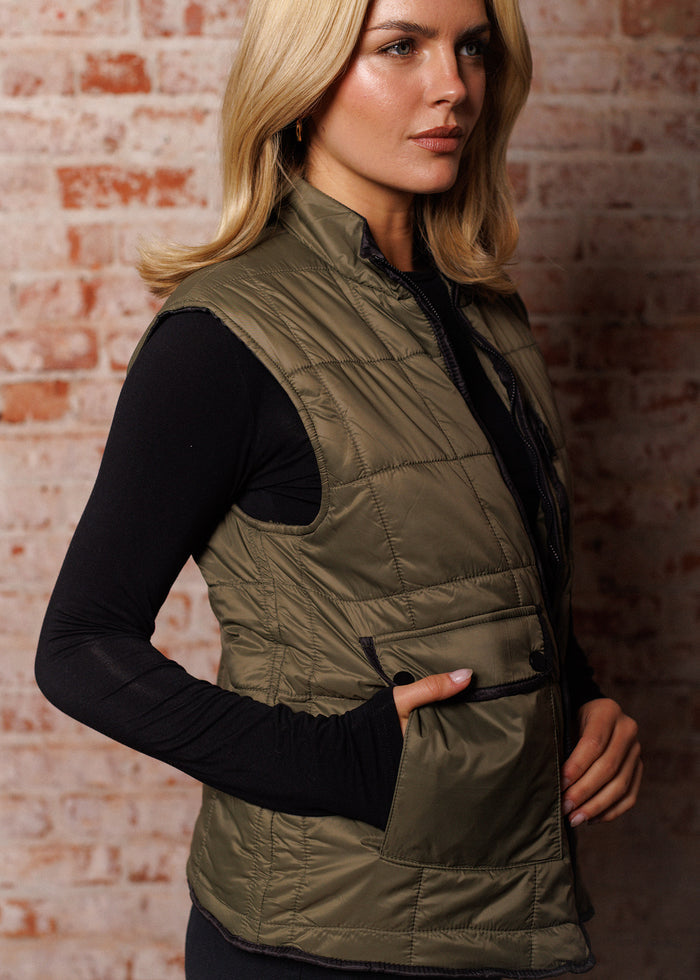 Woman in olive vest and black top stands against a brick wall, gazing to the side, with a hand in her pocket.