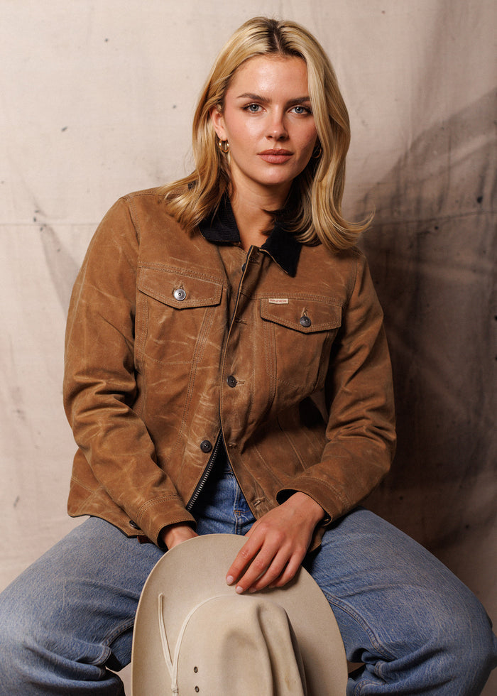Woman in a brown jacket and jeans holds a cowboy hat, sitting against a neutral backdrop, exuding a relaxed, confident vibe.