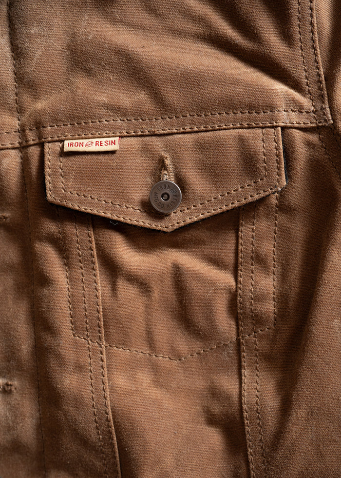 Close-up of a brown fabric jacket pocket with a button and "Iron and Resin" label, highlighting rugged texture and stitching.