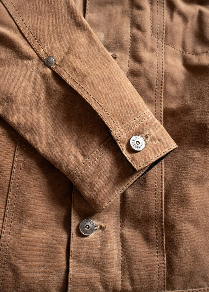 Close-up of a brown canvas jacket sleeve with visible stitching and metal buttons, showcasing durable, rugged fabric texture.