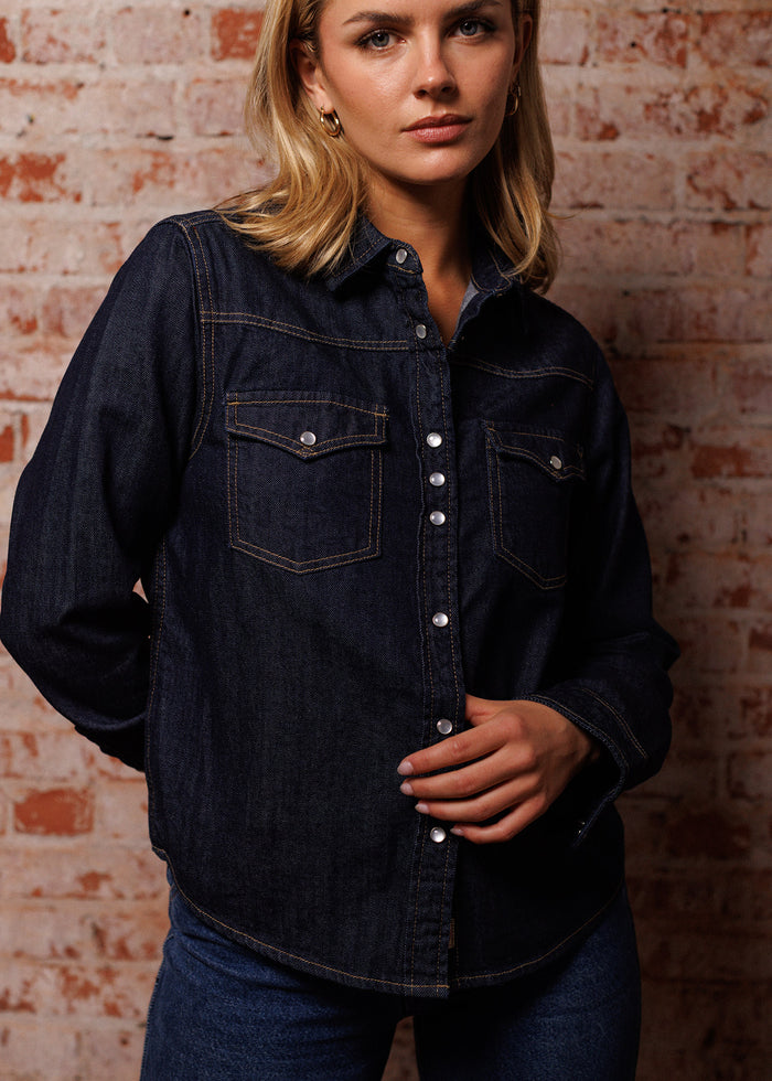A woman in a dark denim shirt stands against a brick wall, looking confidently at the camera with a relaxed pose.