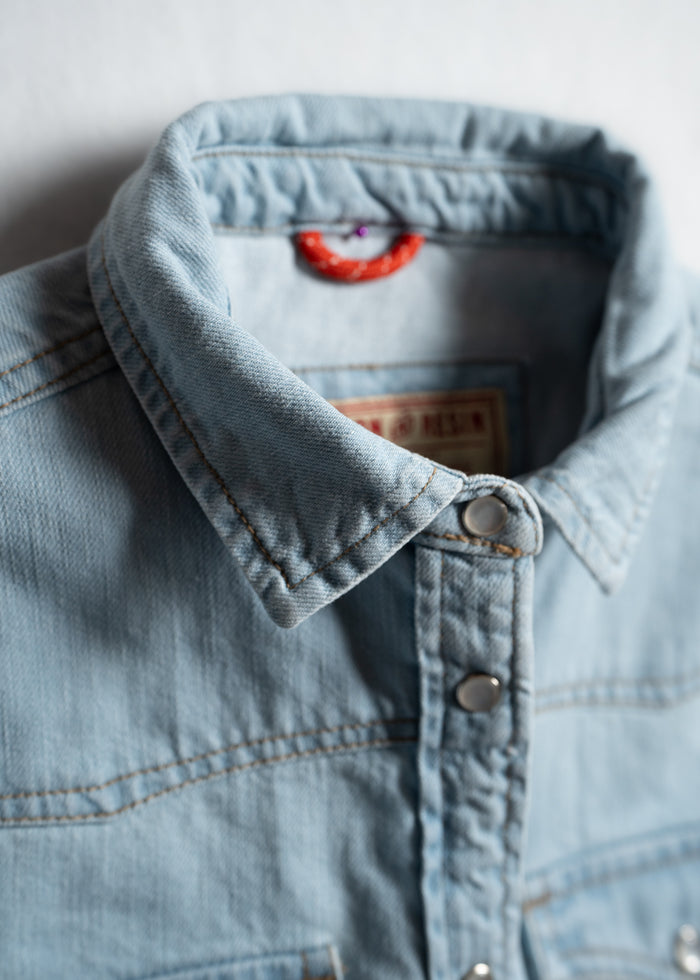 Close-up of a light blue denim shirt collar with metal buttons, highlighting the fabric texture and stitching details.