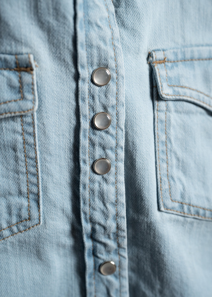 Close-up of a light blue denim shirt with metal snap buttons and visible stitching on two pockets, showcasing texture and design.