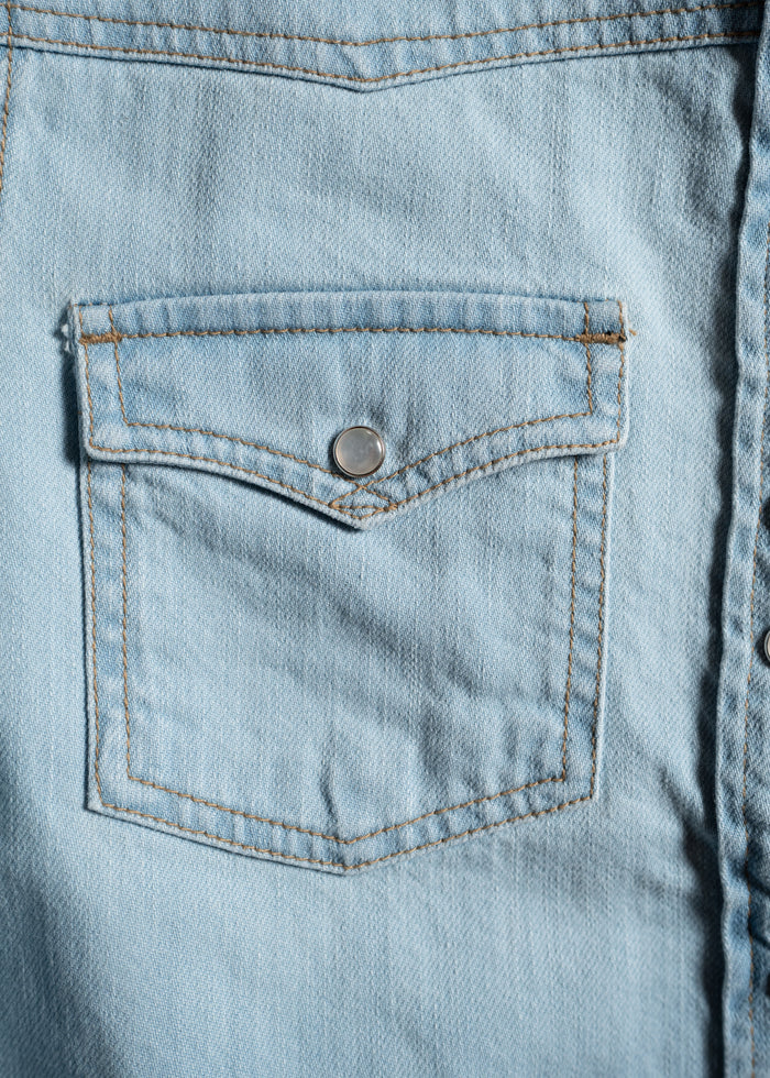 Close-up of a light blue denim shirt pocket with a silver button, showcasing detailed stitching and fabric texture.