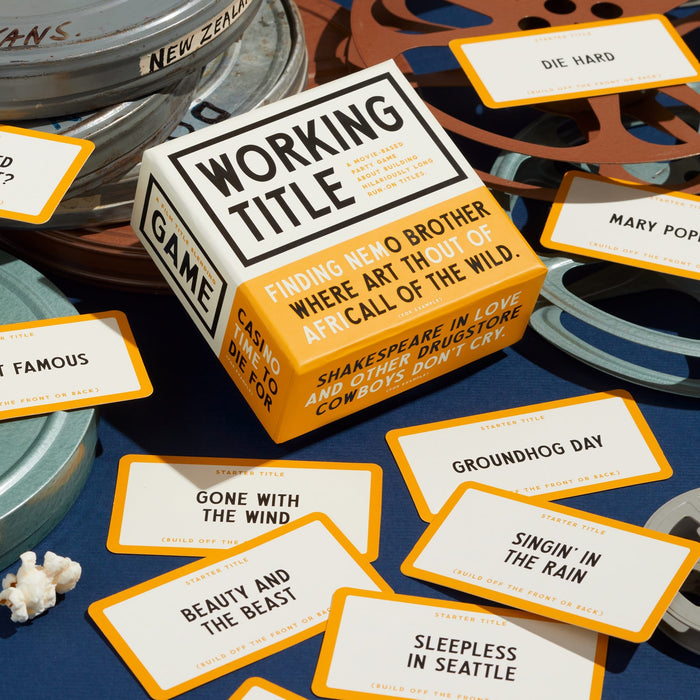 A box of Working Title Game by Brass Monkey sits on a table with movie reel tins, popcorn, and cards from the popular film game featuring titles like Die Hard, Groundhog Day, and Gone with the Wind scattered around.