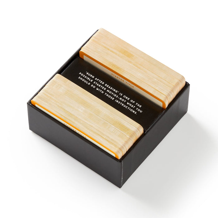 The Working Title Game by Brass Monkey features a black box containing two card stacks with wooden backs and a visible instruction card reading, “Turn after reading. If one of these seems more important, follow those instructions.”.