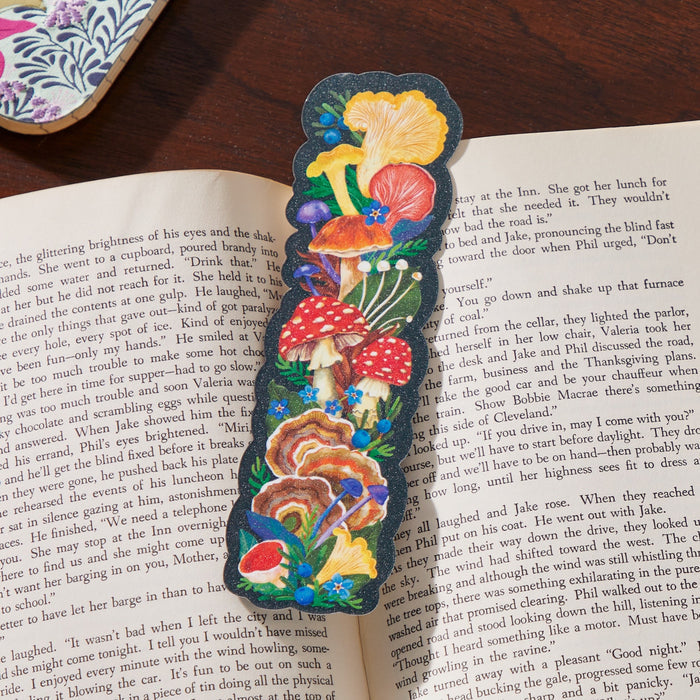 The World of Mushrooms Paper Bookmark by Olga Akbarova showcases vibrant red, yellow, and brown mushrooms with green leaves and blue flowers on a striking black background, adding a colorful touch to any open book.